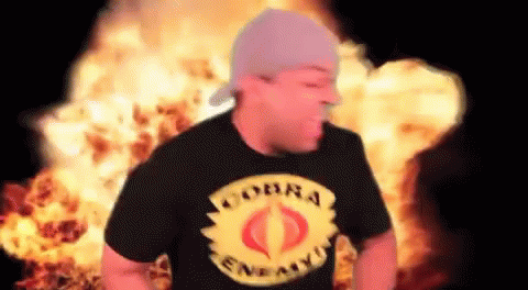 A Man Wearing A Cobra T-Shirt Stands In Front Of A Fire Explosion . GIF