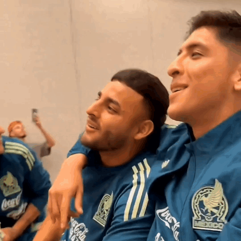 A Man Wearing A Coca Cola Shirt Sits Next To A Man Wearing A Mexico Shirt GIF
