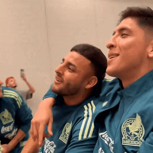 A Man Wearing A Coca Cola Shirt Sits Next To Another Man Wearing A Mexico Shirt GIF