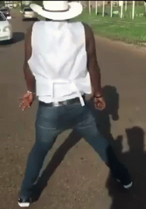 A Man Wearing A Cowboy Hat And A White Vest Is Walking Down The Street GIF