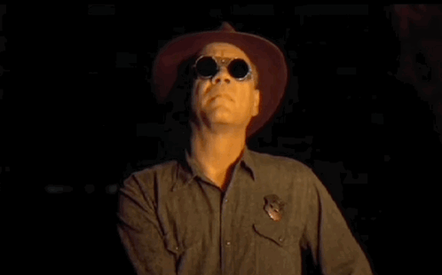 A Man Wearing A Cowboy Hat And Sunglasses Is Standing In The Dark With His Arms Crossed . GIF