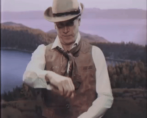 A Man Wearing A Cowboy Hat And Vest Looks At His Watch GIF