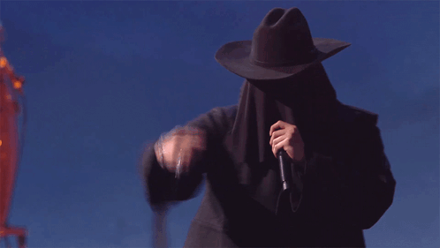 A Man Wearing A Cowboy Hat Covering His Face Holds A Microphone GIF
