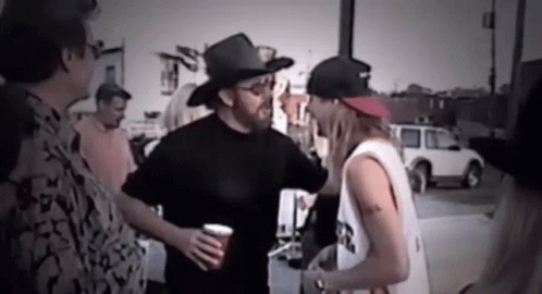 A Man Wearing A Cowboy Hat Is Talking To Another Man While Holding A Cup . GIF