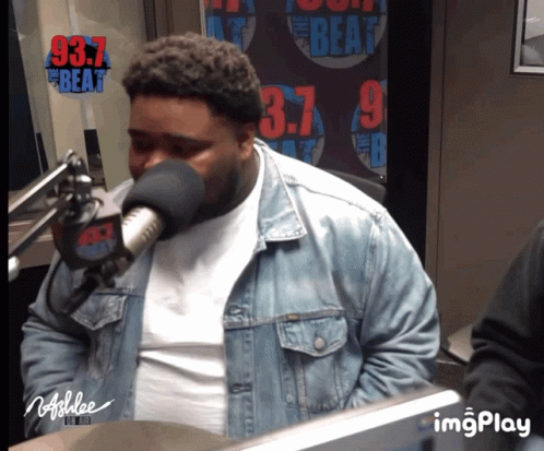 A Man Wearing A Denim Jacket Is Speaking Into A Microphone In Front Of A Sign That Says 97.7 The Beat GIF