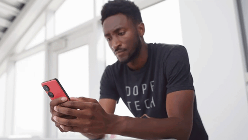 A Man Wearing A Dope Te Ca Shirt Looks At His Cell Phone GIF