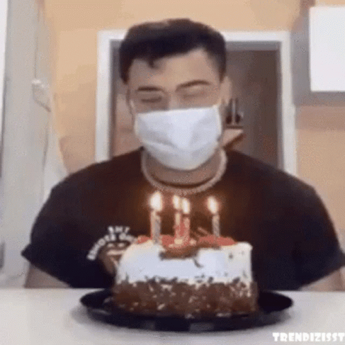 A Man Wearing A Face Mask Is Blowing Out Candles On A Cake . GIF