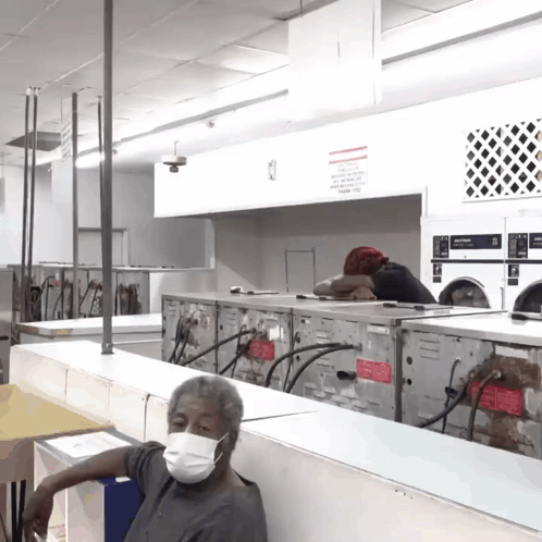 a man wearing a face mask sits in a laundromat surrounded by washers and dryers