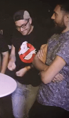 A Man Wearing A Ghostbusters Shirt Is Standing Next To Another Man With His Arms Crossed GIF