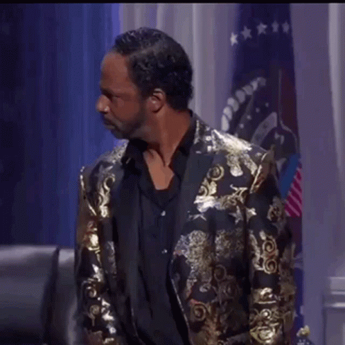 A Man Wearing A Gold Jacket And Black Shirt Is Standing In Front Of A Flag . GIF