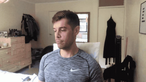 A Man Wearing A Gray Nike Shirt Stands In A Bedroom GIF