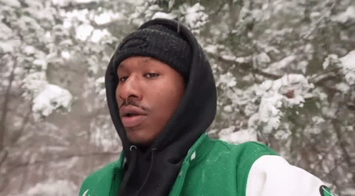 A Man Wearing A Green And Black Jacket And A Black Beanie Is Standing In The Snow . GIF