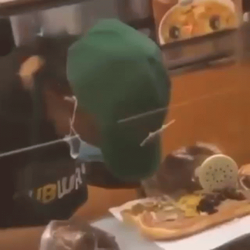 A Man Wearing A Green Hat And A Mask Is Eating A Sandwich . GIF