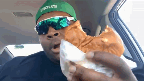 A Man Wearing A Green Hat And Sunglasses Is Eating A Sandwich . GIF