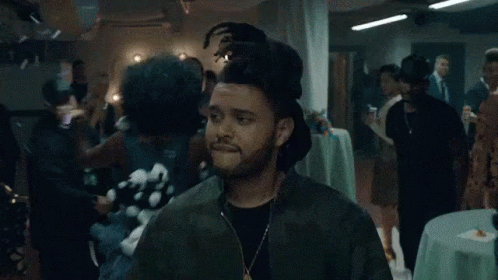 A Man Wearing A Green Jacket And A Necklace Is Standing In A Crowded Room . GIF