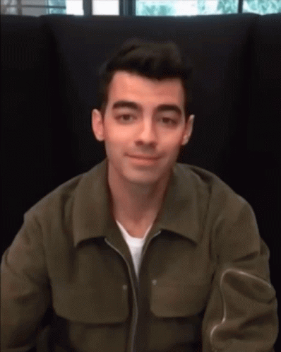 A Man Wearing A Green Jacket Is Sitting In A Chair And Smiling GIF