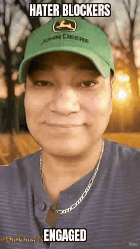 A Man Wearing A Green John Deere Hat And A Necklace Is Smiling And Looking At The Camera . GIF