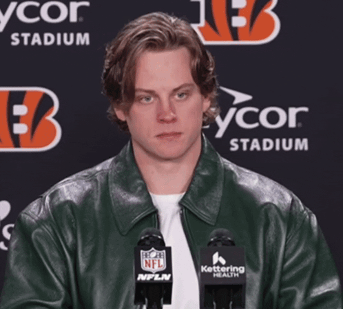A Man Wearing A Green Leather Jacket Stands In Front Of Two Microphones With A Nfl Logo On Them GIF