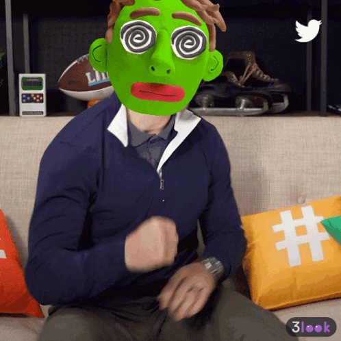A Man Wearing A Green Mask With Hypnotic Eyes Sitting On A Couch GIF