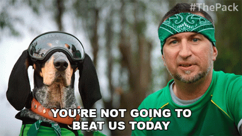 A Man Wearing A Green Shirt And A Bandana Stands Next To A Dog Wearing Goggles And A Sausage GIF