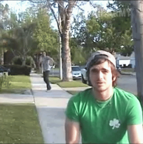 A Man Wearing A Green Shirt With A Shamrock On It Is Standing On A Sidewalk GIF