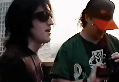 A Man Wearing A Green Shirt With The Letter L On It Stands Next To Another Man Wearing An Orange Hat GIF