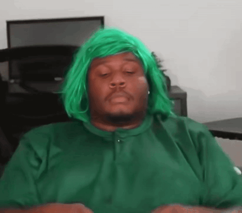 a man wearing a green wig is sitting at a desk with his eyes closed .