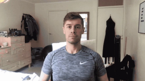 A Man Wearing A Grey Nike Shirt Stands In A Room GIF