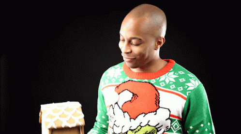 A Man Wearing A Grinch Sweater Looks At A Gingerbread House GIF