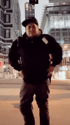 A Man Wearing A Hat And A Black Jacket Stands In Front Of A Tall Building GIF