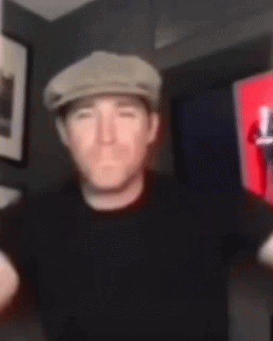 A Man Wearing A Hat And A Black Shirt Is Standing In A Room . GIF