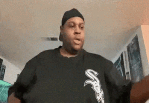 A Man Wearing A Hat And A Black Shirt With The Letter S On It Is Standing In A Living Room . GIF