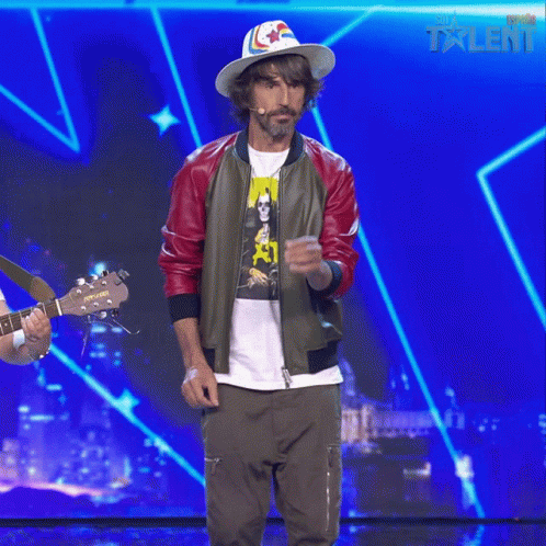 A Man Wearing A Hat And A Bomber Jacket Stands On A Stage With A Blue Background That Says Talent GIF