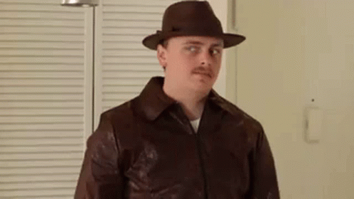 A Man Wearing A Hat And A Brown Leather Jacket Is Standing In Front Of A Window . GIF
