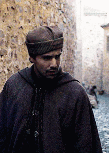 A Man Wearing A Hat And A Cape Is Walking Down A Narrow Street GIF