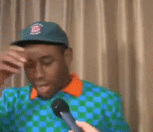 A Man Wearing A Hat And A Checkered Shirt Is Covering His Face With His Hand GIF