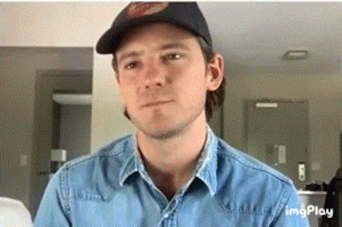 A Man Wearing A Hat And A Denim Shirt Looks At The Camera GIF