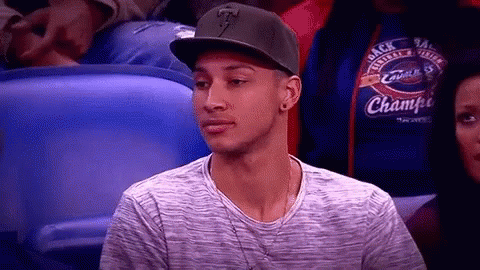 A Man Wearing A Hat And A Gray Shirt Is Sitting In A Stadium Watching A Game . GIF