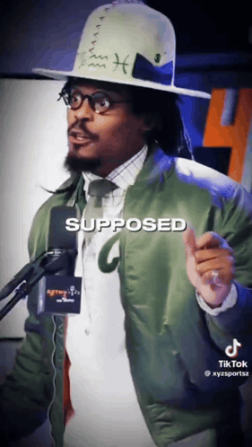 A Man Wearing A Hat And A Green Jacket Is Standing In Front Of A Microphone With The Word Supposed Written On It GIF
