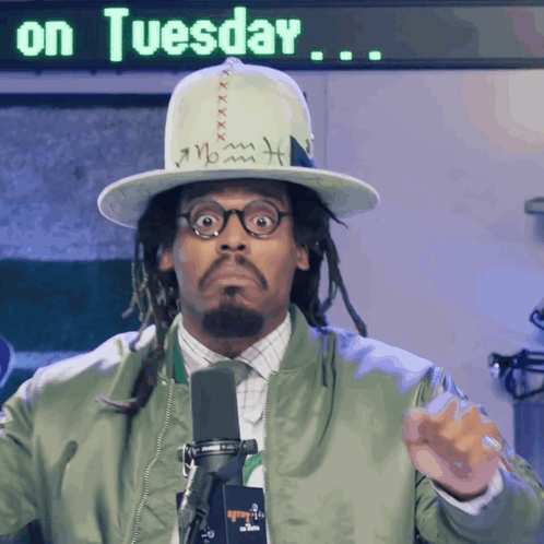 A Man Wearing A Hat And A Green Jacket Stands In Front Of A Sign That Says " On Tuesday " GIF