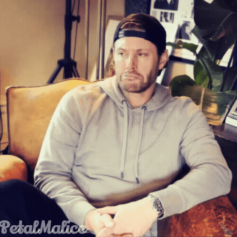 A Man Wearing A Hat And A Grey Hoodie Is Sitting On A Couch With Petal Malice Written On The Bottom GIF