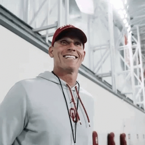 A Man Wearing A Hat And A Hoodie Is Smiling While Standing In A Gym . GIF