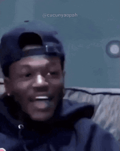 A Man Wearing A Hat And A Jacket Is Sitting On A Couch And Smiling . GIF