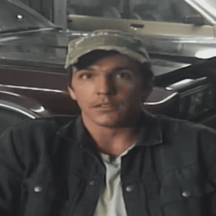 a man wearing a hat and a jacket stands in front of a parked car