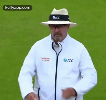 A Man Wearing A Hat And A Jacket That Says Emirates Fly Better Is Standing On A Field . GIF