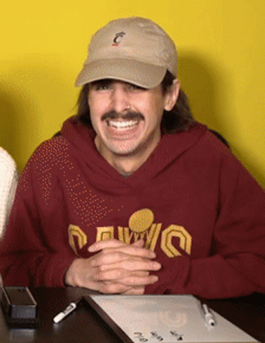 A Man Wearing A Hat And A Maroon Sweatshirt That Says ' Cleveland ' On It Sits At A Desk GIF