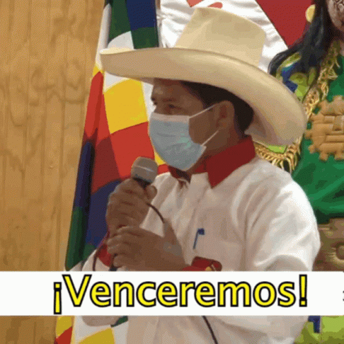 A Man Wearing A Hat And A Mask Holds A Microphone In Front Of A Banner That Says Venceremos GIF