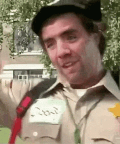 A Man Wearing A Hat And A Name Tag Is Smiling And Looking At The Camera . GIF