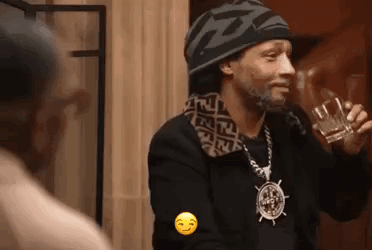 A Man Wearing A Hat And A Necklace Is Holding A Glass Of Alcohol . GIF