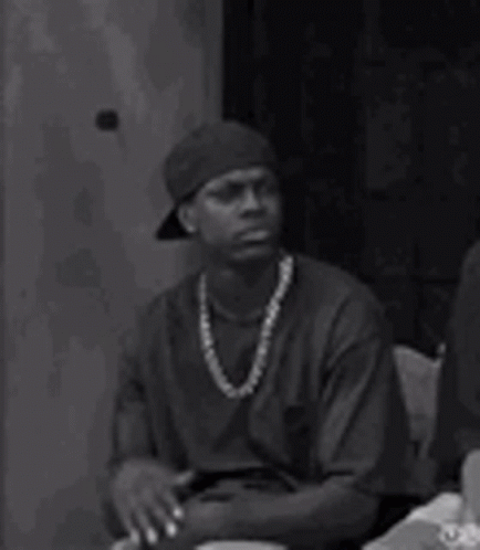 A Man Wearing A Hat And A Necklace Is Sitting At A Table With His Hands Folded . GIF